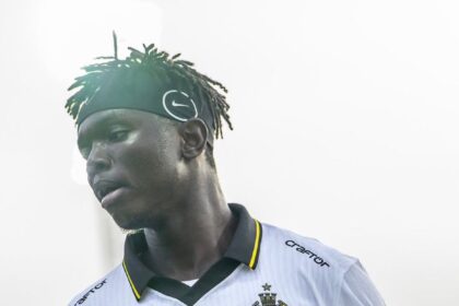 Soccer player in a white jersey with black/yellow trim, holding a soccer ball under his right arm.