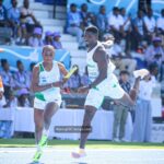 Two sprinters in white and green uniforms pass a baton during a relay race on a blue track with a cheering crowd in the stands behind them  trybalnews