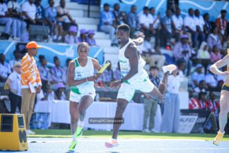 Two sprinters in white-and-green uniforms pass a baton during a relay race on a blue track, with a cheering crowd in the stands behind them.