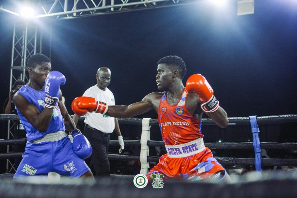 WhatsApp Image 20260504 at 190439 1 Trybal News Two male boxers exchange punches in a ring one in blue trunks and gloves and the other in red with Delta State branding as a referee watches from behind trybalnews