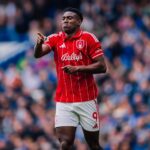Soccer player in a red striped jersey points forward during a match with a blurred crowd in the stands behind him  trybalnews