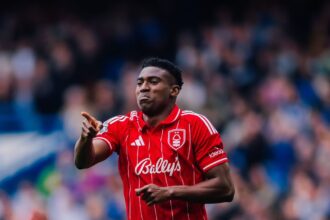 Soccer player in a red striped jersey points forward during a match with a blurred crowd in the stands behind him.