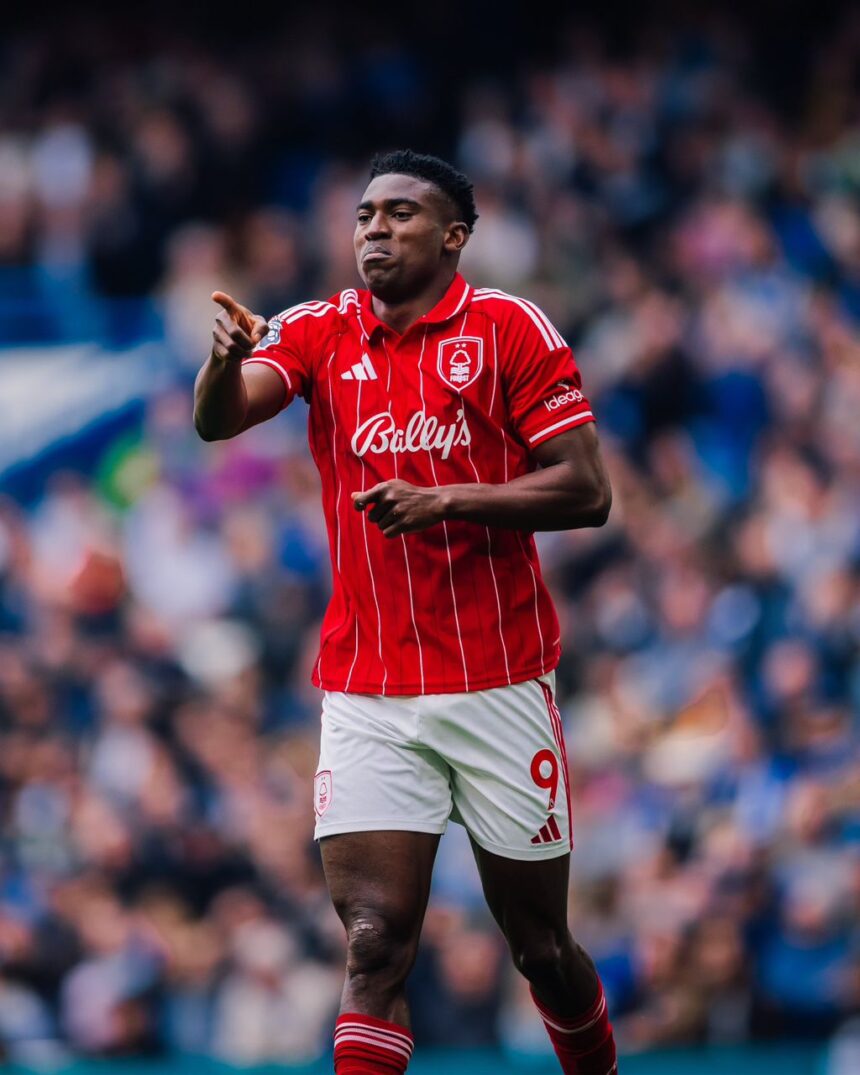 Soccer player in a red striped jersey points forward during a match with a blurred crowd in the stands behind him  trybalnews