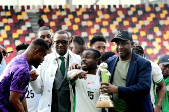 Group of soccer players posing with a trophy on the field after a match, stadium seating in multicolored background.