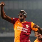 Football player in orange red Galatasaray jersey raises fist in celebration wearing protective goggles stadium backdrop behind  trybalnews