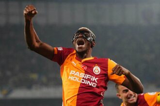 Football player in orange-red Galatasaray jersey raises fist in celebration, wearing protective goggles; stadium backdrop behind.