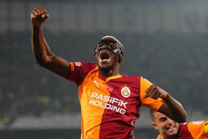 Football player in orange-red Galatasaray jersey raises fist in celebration, wearing protective goggles; stadium backdrop behind.