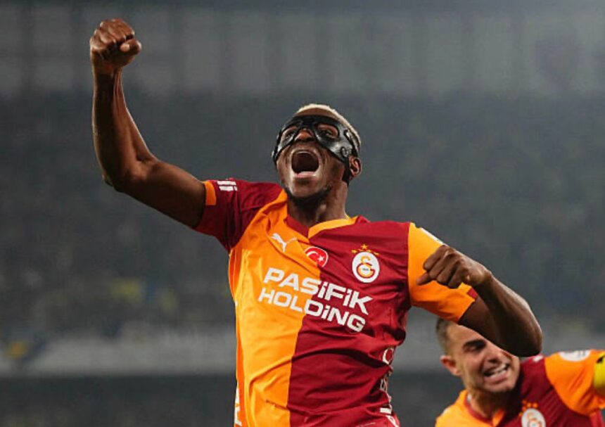 Football player in orange red Galatasaray jersey raises fist in celebration wearing protective goggles stadium backdrop behind  trybalnews