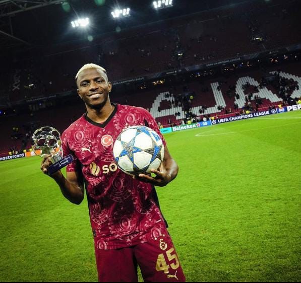 WhatsApp Image 20260508 at 075340 Trybal News Soccer player in maroon kit holding a trophy and a star patterned ball on a well lit stadium pitch with empty stands in the background trybalnews