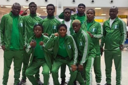 Group of athletes in matching green tracksuits posing for a team photo in an indoor facility, some crouching in front and others standing behind. The uniforms feature Nigeria flag patches on the jackets.