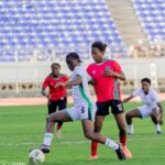 Soccer match in progress with players in white and red uniforms contesting the ball on a green field blue stadium seats in the background  trybalnews