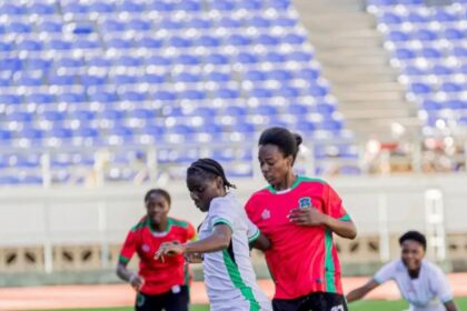 Soccer match in progress with players in white and red uniforms contesting the ball on a green field; blue stadium seats in the background.