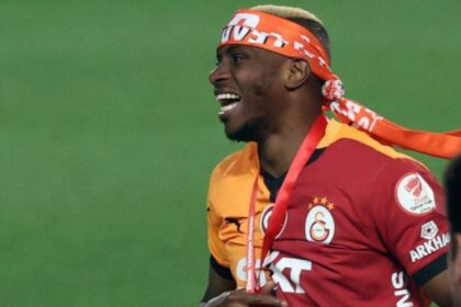 Soccer player in a red-orange Galatasaray kit with a headband, smiling on the field during a celebration.