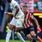 Two soccer players contest a ball on the field a white kit player shields the ball while a red and white striped opponent closes in  trybalnews