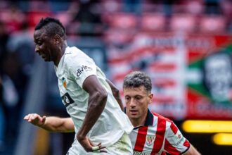 Two soccer players contest a ball on the field; a white-kit player shields the ball while a red-and-white striped opponent closes in.