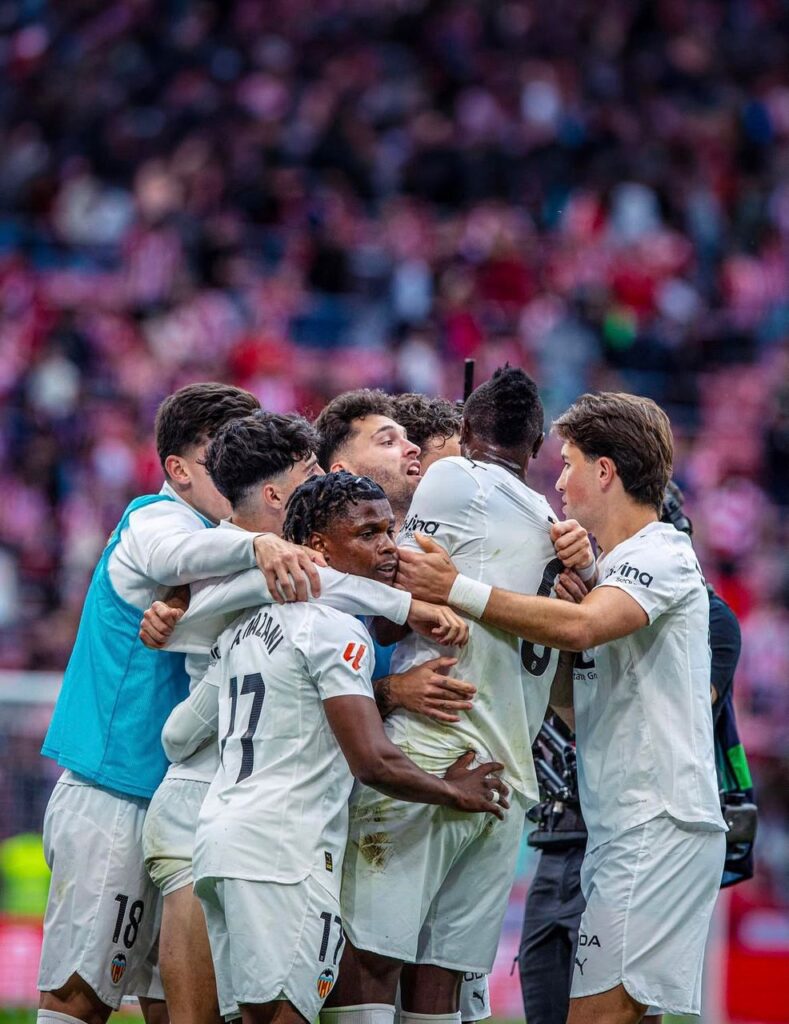 WhatsApp Image 20260510 at 200813 Trybal News A group of soccer players in white uniforms celebrate with a group hug on the field after a goal with a blurred crowd in the stands behind them trybalnews