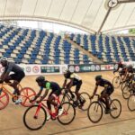 Group of cyclists racing on an indoor velodrome track under a curved roof with colorful bikes and helmets in a pack  trybalnews