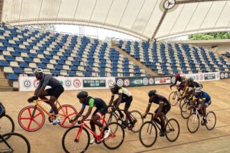 Group of cyclists racing on an indoor velodrome track under a curved roof, with colorful bikes and helmets in a pack.