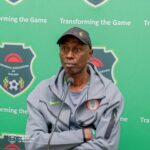 Man wearing a gray jacket and cap sits at a microphone at a Malawi Football Association press conference with a green backdrop  trybalnews