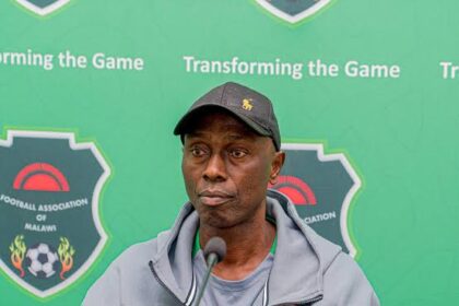 Man wearing a gray jacket and cap sits at a microphone at a Malawi Football Association press conference with a green backdrop