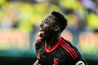 Soccer player in a black jersey with red trim celebrates a goal, tongue out, running on the field with a blurred stadium background