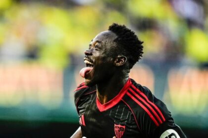 Soccer player in a black jersey with red trim celebrates a goal, tongue out, running on the field with a blurred stadium background