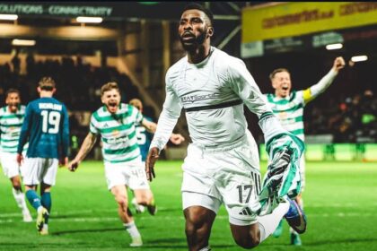 Soccer player in a white kit (number 17) runs toward the camera as teammates celebrate in the background on a green pitch.