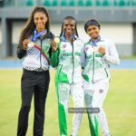 Three female athletes stand together on a track field smiling and showing their medals in team uniforms  trybalnews