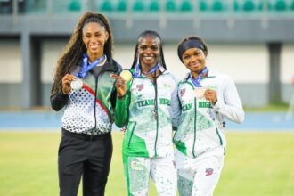 Three female athletes stand together on a track field, smiling and showing their medals in team uniforms.