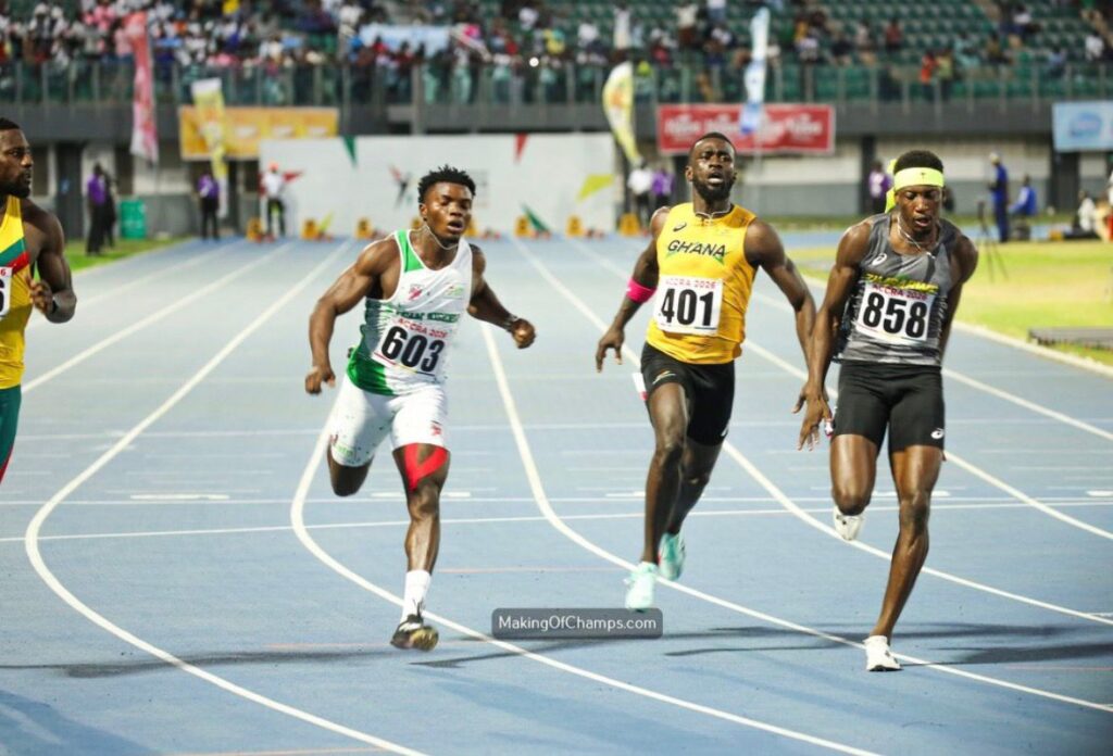 Three male sprinters race down a blue track each wearing bibs 603 401 858 as crowds watch from the stands one runner wears a Ghana jersey while another wears green white green and a third wears black with a white headband  trybalnews