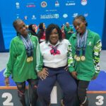 Three people posing on a podium a seated woman in a white shirt and scarf flanked by two young athletes in green tracksuits wearing medals with a blue competition backdrop and other people in the background  trybalnews