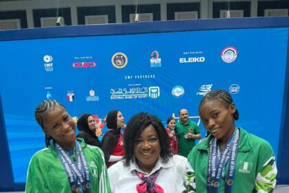 Three people posing on a podium: a seated woman in a white shirt and scarf flanked by two young athletes in green tracksuits wearing medals, with a blue competition backdrop and other people in the background.