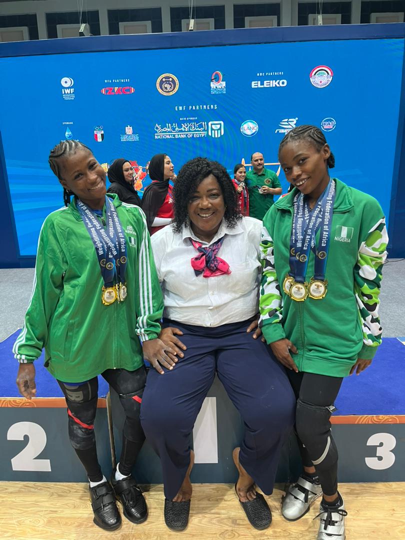Three people posing on a podium a seated woman in a white shirt and scarf flanked by two young athletes in green tracksuits wearing medals with a blue competition backdrop and other people in the background  trybalnews