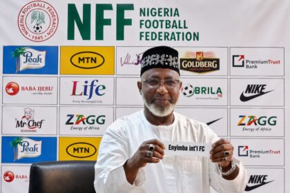 Nigerian football official in white attire holding a sign that says 'Enyimba int'l FC' at a press conference with NFF backdrop behind him.