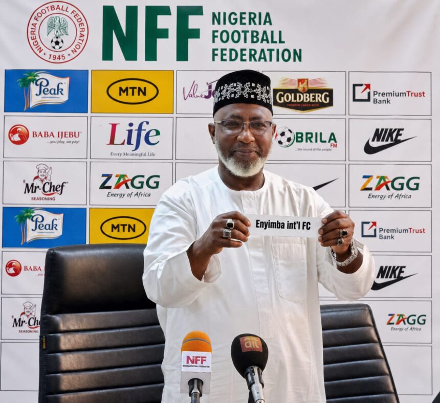 Nigerian football official in white attire holding a sign that says Enyimba intl FC at a press conference with NFF backdrop behind him  trybalnews