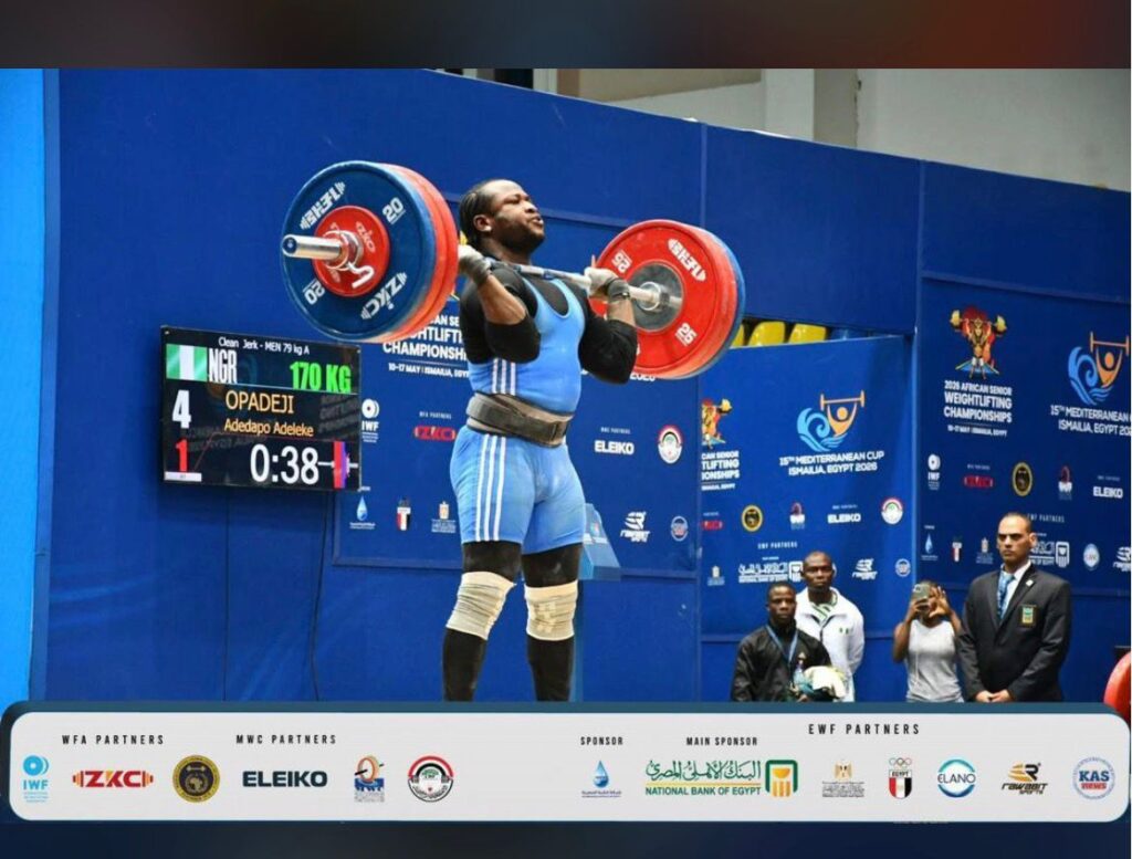 Male weightlifter in a blue singlet lifts a heavy barbell overhead during a competition with a scoreboard and blue competition venue in the background  trybalnews