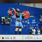 Athlete lifts a loaded barbell overhead on a blue competition stage scoreboard visible on the left and officials in the background  trybalnews