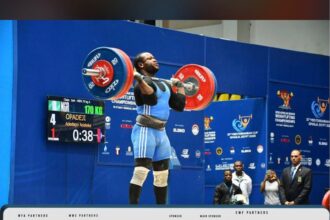 Athlete lifts a loaded barbell overhead on a blue competition stage, scoreboard visible on the left and officials in the background.