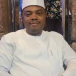 Man wearing a white traditional outfit and cap seated and smiling at the camera in a dining setting  trybalnews
