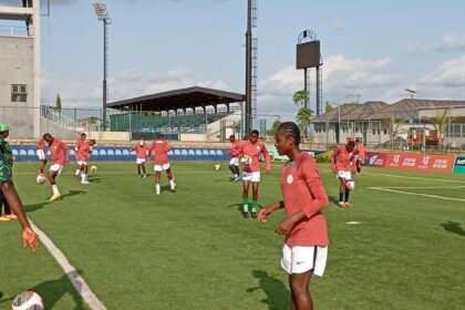 Soccer training on a sunny field with female players in red jerseys and white shorts handling balls statuesque on green turf.