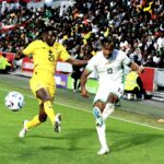 Two soccer players in yellow and white kits chase a ball on a green pitch with a crowded stadium in the background mid action  trybalnews