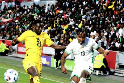 Two soccer players in yellow and white kits chase a ball on a green pitch with a crowded stadium in the background, mid-action.