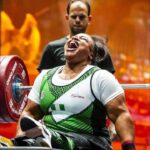 Female powerlifter in a wheelchair with a green and white singlet shouting with effort as she lifts a heavy barbell spotter behind her in a gym  trybalnews