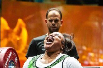 Female powerlifter in a wheelchair with a green and white singlet shouting with effort as she lifts a heavy barbell; spotter behind her in a gym.