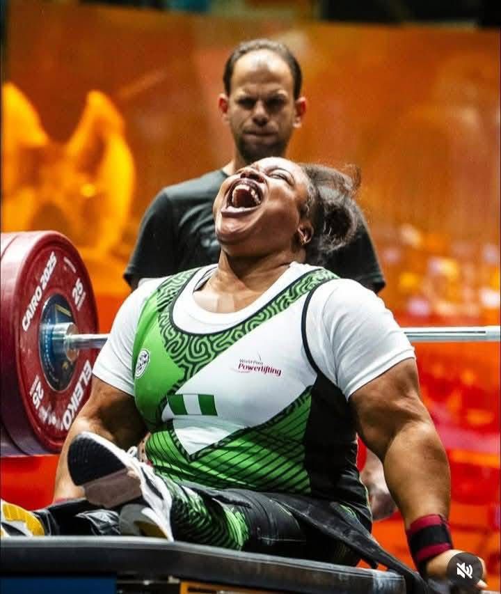 Female powerlifter in a wheelchair with a green and white singlet shouting with effort as she lifts a heavy barbell spotter behind her in a gym  trybalnews