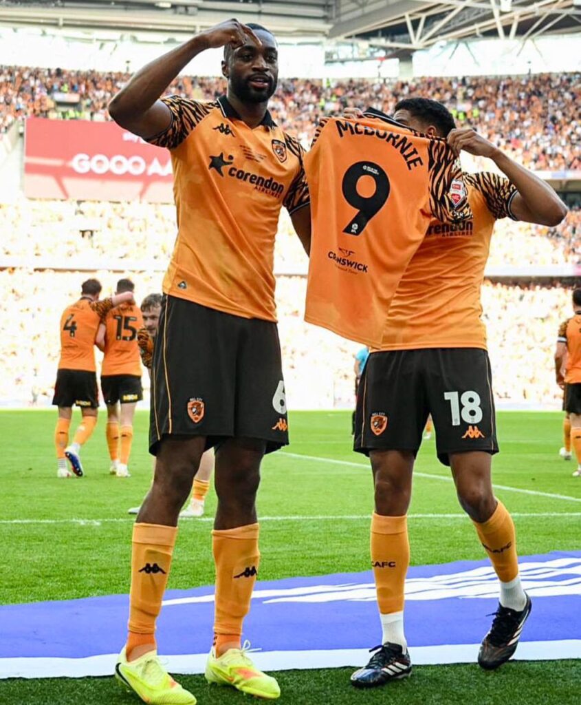 WhatsApp Image 20260523 at 184629 Trybal News Two soccer players in orange kits celebrate on the pitch lifting their jerseys in a stadium crowd trybalnews