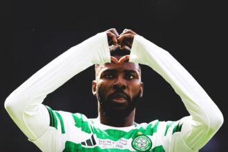 Soccer player in a green-and-white hooped jersey forms a heart shape with his arms during a celebration on the field