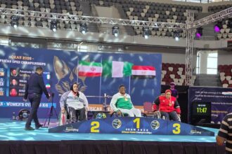 Three wheelchair athletes stand on a podium at the African Open Championships, with blue banners, flags, and tiered placements behind them.