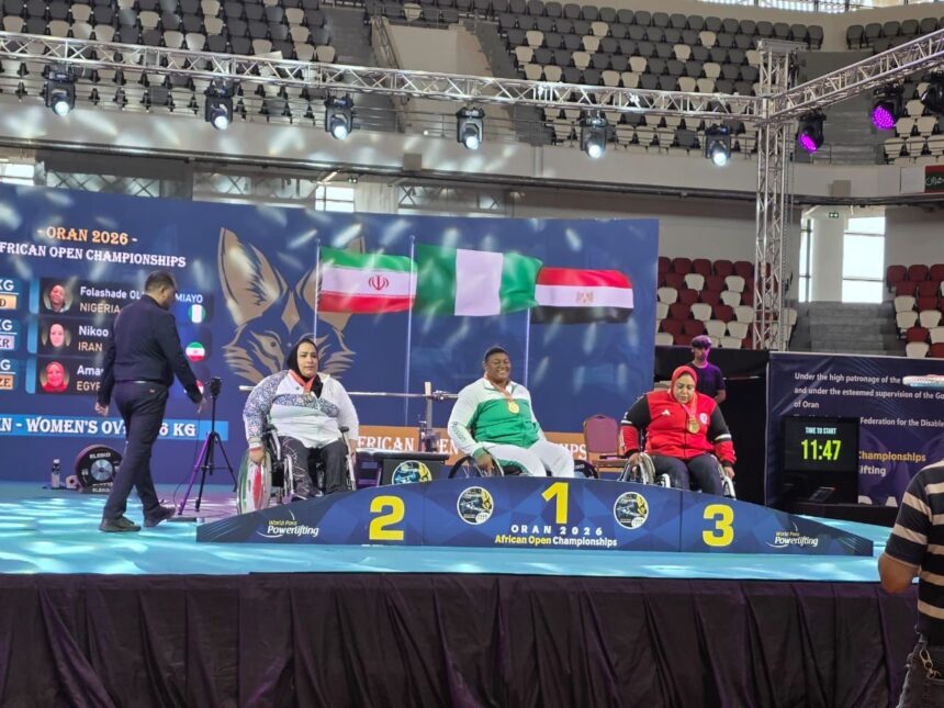 Three wheelchair athletes stand on a podium at the African Open Championships with blue banners flags and tiered placements behind them  trybalnews