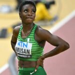 Female sprinter in a green striped uniform with bib reading AMUSAN and Budapest 23 standing with hands on hips on a track looking upward  trybalnews